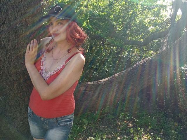 YanaFiesta mit roten Haaren, die ein rotes Tanktop mit Spitzenbesatz, blaue Jeans und eine schwarze Mütze trägt, steht neben einem Baum und macht ein verspieltes Gesicht mit einem Augenzwinkern, wobei Sonnenstrahlen zu sehen sind..