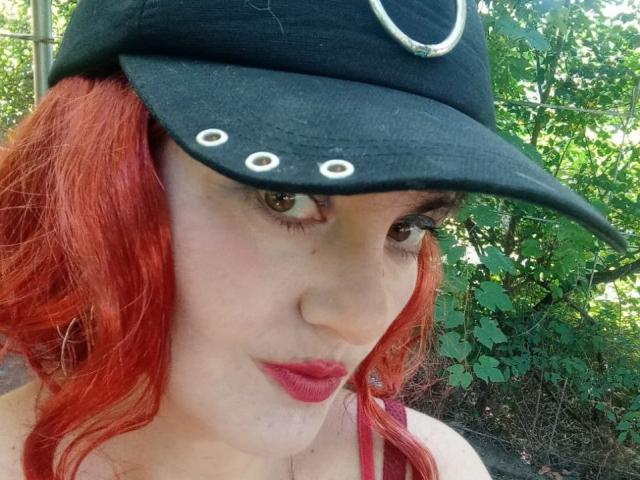 YanaFiesta mit roten Haaren, die eine schwarze Mütze mit weißen Ösen trägt, hat helle Haut, grüne Augen und roten Lippenstift. Ihre Mütze ist leicht schief. Sie ist draußen..