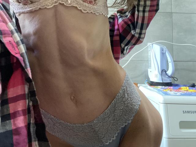 Kabblasha's slim, toned torso with visible ribs, wearing lace underwear and a pink bra, has a plaid shirt hanging off. Sheer white electric kettle on the right.