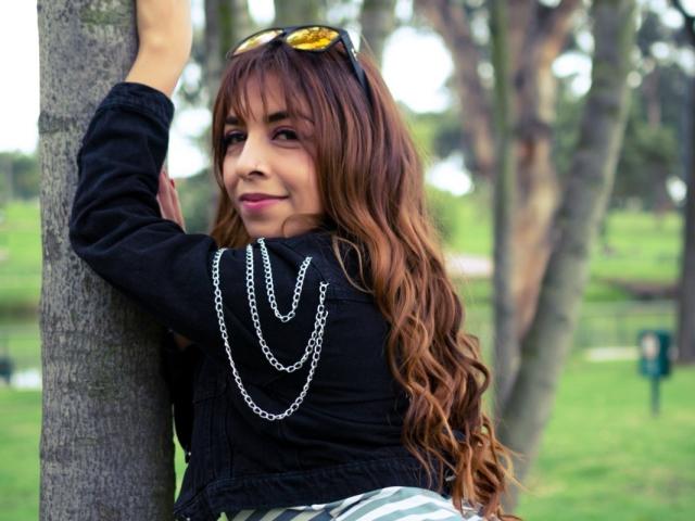 AmlieferDerline with long wavy brown hair, black sweater, and chain necklace, leaning on a tree, wearing sunglasses on head, smiling, outdoors, green background.