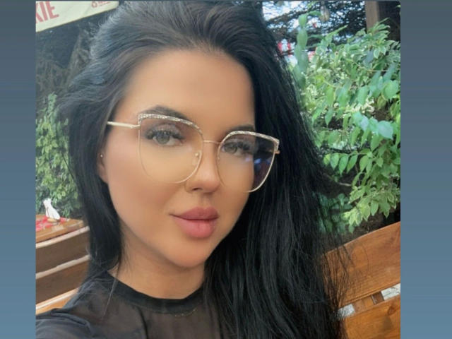 HeisseVerra-hot with long black hair, wearing clear glasses with sparkly edges, black sheer top, and pink lips, sitting outdoors, looking at camera with a slight smile.