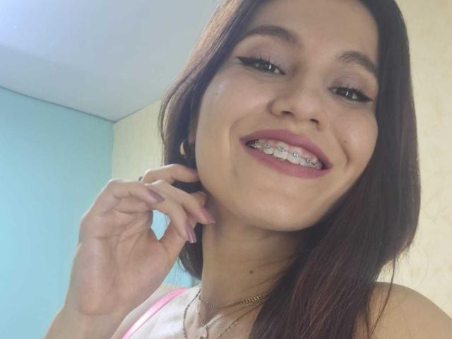 AmallaVega's smiling face with straight brown hair, dark pink lipstick, and metal teeth, holding her chin with a hand, wearing a gold necklace, and a white top.