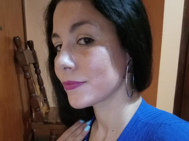 Satika with black hair, blue eyes, pink lipstick, and blue shirt, wearing large hoop earrings, has shiny skin and light blue nails, standing near wooden chairs.