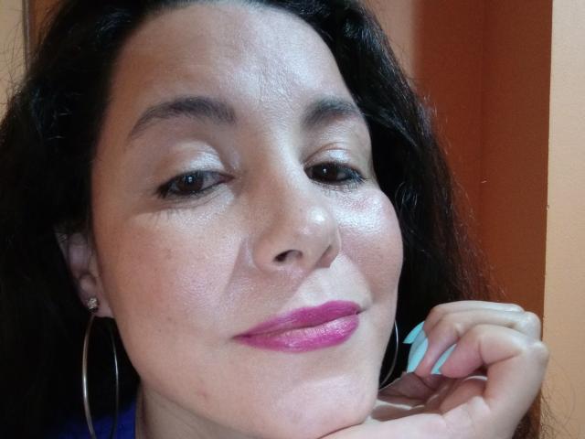 Satika's face, brown eyes, pink lipstick, slightly flushed skin, long dark hair, hoop earrings, light blue nails, resting chin on hand, subtle smile, medium skin tone, relaxed expression.