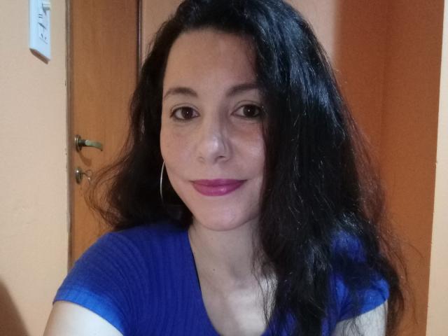 Satika with long black wavy hair, wearing a blue V-neck shirt, pink lipstick, and hoop earrings, smiling at the camera. Her tits are small and her ass is unseen.
