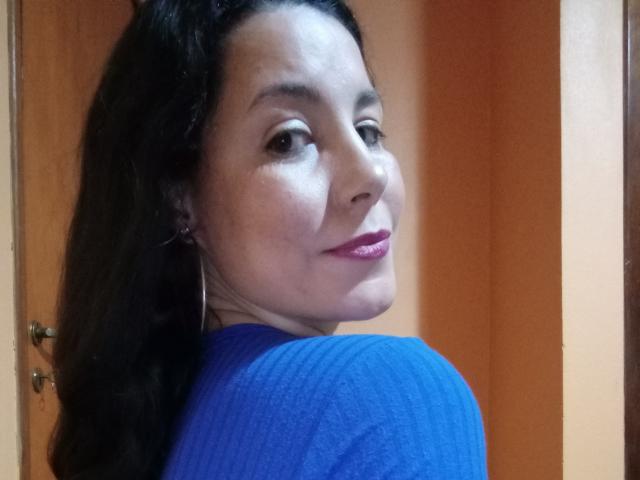 Satika with wavy black hair, blue ribbed shirt, pink lipstick, and hoop earrings, looking over her shoulder, smiling slightly. Her ass is partially visible.