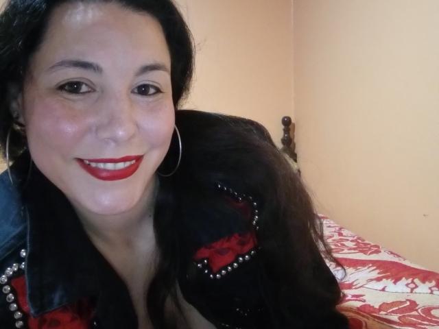 Satika with red lipstick, black hair, and hoop earrings, wearing a black leather jacket with red and black fabric, smiling at the camera, showing her tits.