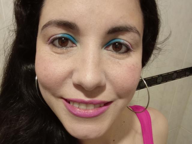 Satika with blue eye makeup, pink lipstick, and hoop earrings, smiling at the camera. She has dark hair, brown eyes, and is wearing a pink top.