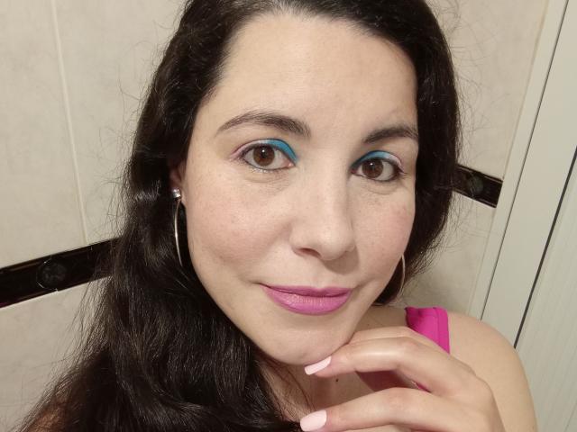 Satika with dark hair, blue eyeshadow, pink lipstick, pink nails, pink top, small hoop earrings, brown eyes, smiling, hand near chin, fair skin, slight freckles.