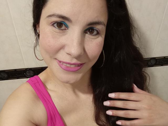 Satika with pink lipstick, blue eyeshadow, and hoop earrings, smiling at the camera, wearing a pink tank top, touching her long dark hair, showing her pink-nailed hand.