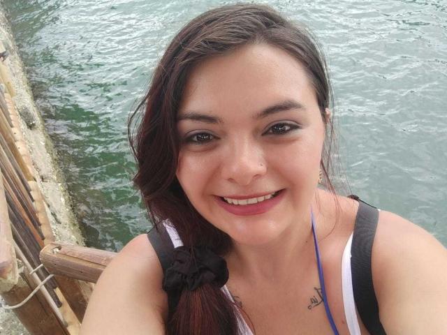 KatherinLatin ha i capelli castano scuro raccolti in una treccia, un elastico nero per capelli e una canotta bianca, sorride con il rossetto rosa, mostrando un piccolo tatuaggio sul petto..