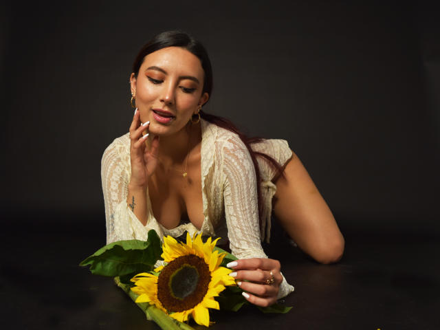CarlaFranco, met lang donker haar, draagt een kanttop en ligt op een zwarte ondergrond. Ze raakt met haar rechterhand de bloemblaadjes van een zonnebloem aan, toont haar borsten, en glimlacht. De achtergrond is zwart..