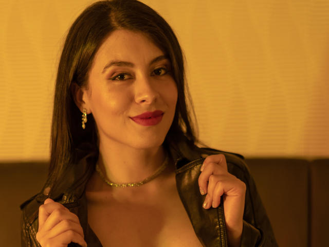 JaneLondon with dark brown hair, red lipstick, and a black leather jacket, holding it open to show her tits, wearing a necklace and earrings, smiling warmly.