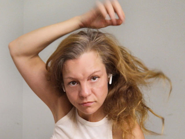 HotLena1984-sex with long, wavy blonde hair, wearing a white sleeveless top, has her arm raised, looking serious, and wearing white earbuds. Her tits are not visible.
