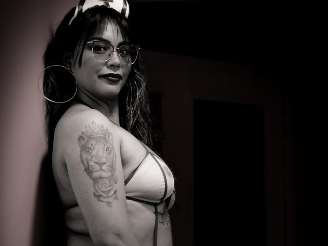 VeronicaSaienz with glasses, dark lipstick, and hoop earrings, wearing a strappy top, showing a tiger rose tattoo on her right arm, in a black-and-white photo.