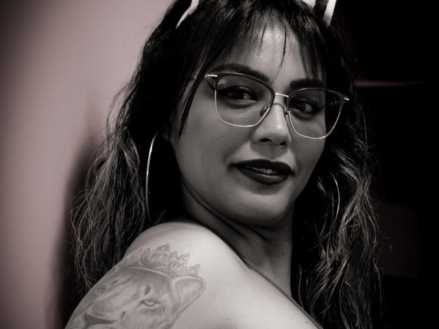 VeronicaSaienz with glasses, dark lipstick, wavy hair, hoop earrings, and a tattoo of a lion's face on her shoulder. Black-and-white, slightly grimy, confident smirk.