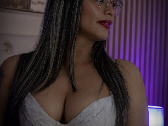 VeronicaSaienz with long black hair, glasses, and red lipstick, wearing a white lace bustier that reveals her large, round tits. Dim purple light in background.