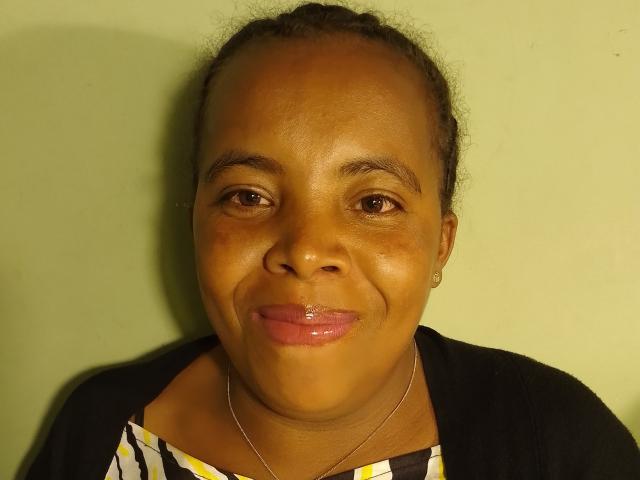 ElysiaX has dark brown skin, small earrings, a silver necklace, and is wearing a black cardigan over a yellow-white-striped top, with shiny pink lips and a slight smile.