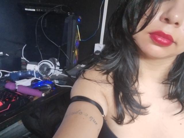 YosefinFabouls has black wavy hair, red lipstick, and a tattoo on her right arm. She's wearing a black strap, and a pink vibrator is on a desk.