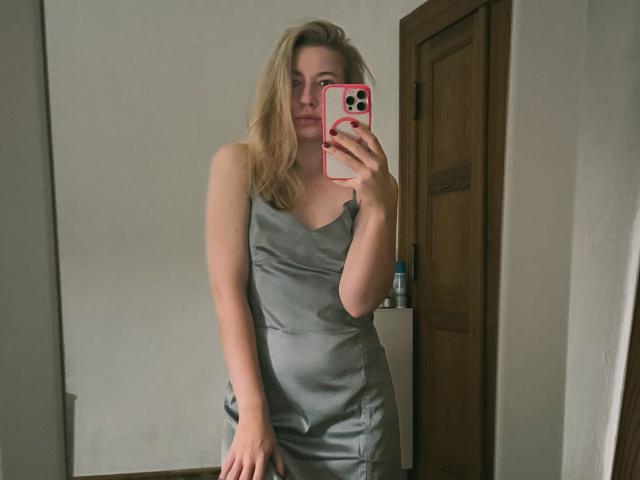 MiaFlirt, con cabello rubio, vestida con un vestido gris, sin mangas y ajustado, se toma un selfie frente al espejo con un teléfono rosa, de pie en una habitación con una puerta de madera..