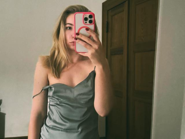 MiaFlirt with blonde hair, wearing a green satin dress, holding a pink phone, showing her tits, mirror selfie, wooden door in background, red nail polish, subtle smile.