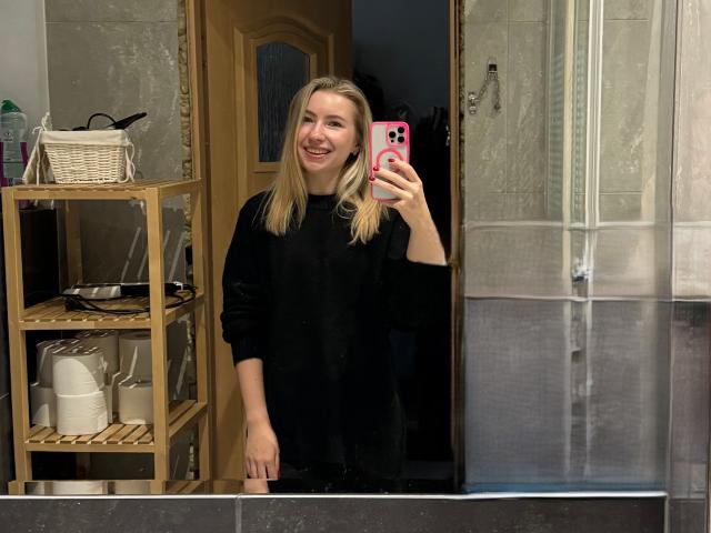 MiaFlirt, com cabelo loiro, sorrindo, vestindo um moletom preto, segurando um telefone rosa, tirando uma selfie no espelho em um banheiro, mostrando seu rosto fofo e sorriso sedutor..