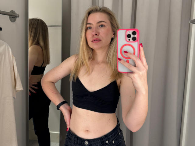 MiaFlirt, con cabello rubio, vistiendo un top deportivo negro, mostrando sus pechos, y jeans oscuros, tomando una selfie frente al espejo con un teléfono rosa, en un baño..