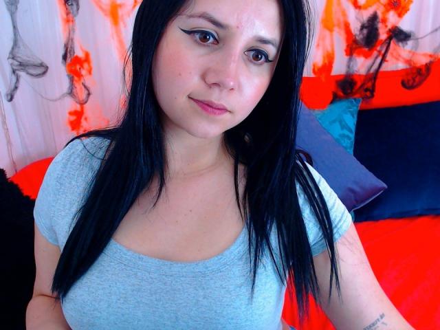 Kkatalina with black hair, blue eyes, and light skin, wearing a gray tee, showing small tits, and a subtle smile, with a tattoo on her right arm.