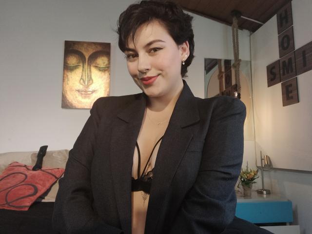 CristineBonnye with short black hair, wearing a black blazer, exposing black lace bra, showing small tits, and red lipstick, smiling in a room with wall art.