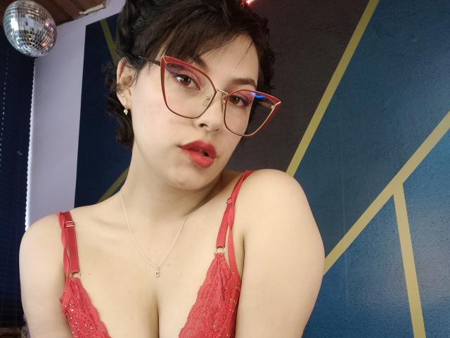 CristineBonnye with red glasses, red lace bra, and red lipstick, showing her tits and a small nose ring, against a blue and yellow wall with a mirror.
