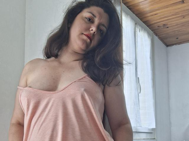 LaFrancaiseJessy com seios médios, blusa rosa com um ombro descoberto, cabelo castanho ondulado, batom vermelho, em pé em um quarto iluminado pelo sol, teto de madeira, cortinas brancas, cabeça ligeiramente inclinada, expressão confiante..