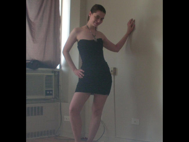 Chiara177-hot, standing, wearing a black strapless mini-dress, showing her ass, with her hand on the wall, smiling, brown hair in a ponytail, necklace, in a dimly lit room.