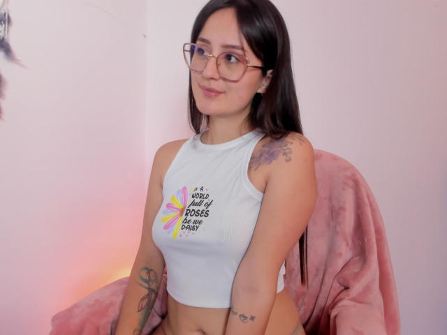 NinaBryons with long black hair, glasses, white crop top, tattoos on arms, pink chair, pink blanket, soft smile, cute, medium tits, small ass, light skin, pink lighting.