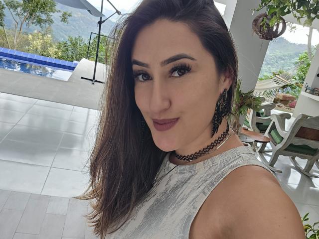 HotCarolina69 with medium tits, wearing a gray patterned top, black choker, and earrings, has brown hair, dark makeup, and a subtle smile, standing by a pool.