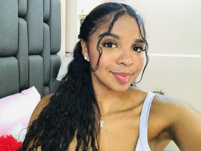 LisaMendez has long black curly hair, dark brown eyes, and pink lipstick. She's wearing a white tank top, showing her small tits and armpit hair. She's smiling.