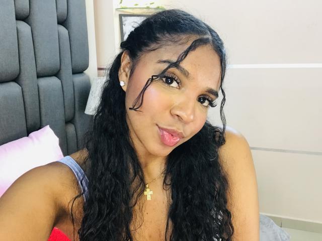 LisaMendez with dark curly hair, sticking out her tongue, wearing a necklace, and showing off her small tits in a pink top, sitting on a bed.