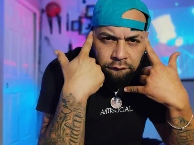 ValsyHot with a teal backwards cap, black t-shirt, tattoos on arms, and a beard, pointing fingers at his face, wearing a silver necklace with a circular pendant.