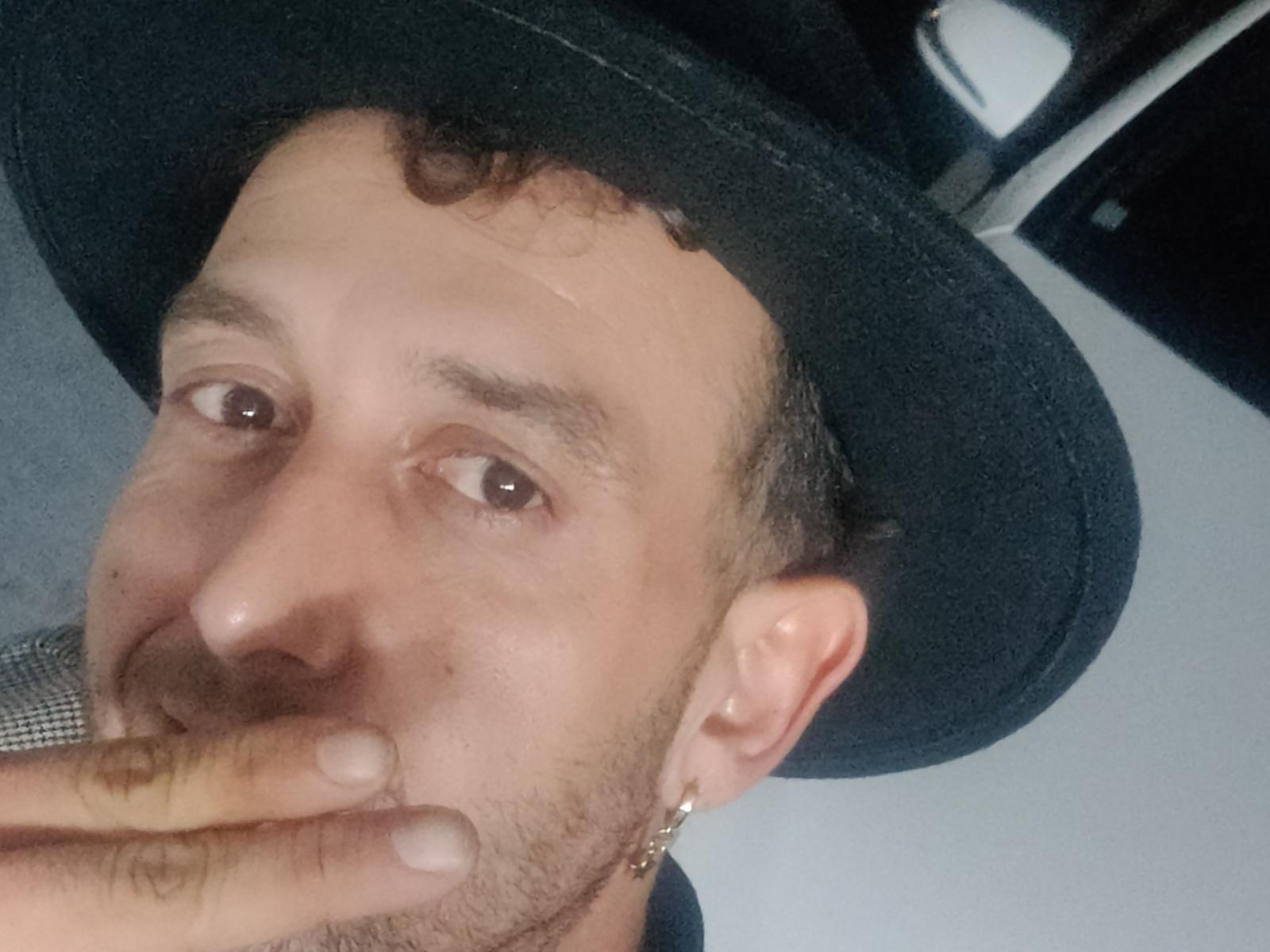 SexyBigH with a black hat, curly brown hair, light skin, brown eyes, and a gold earring, covering his mouth with his hand, showing a slight scar above his eye.