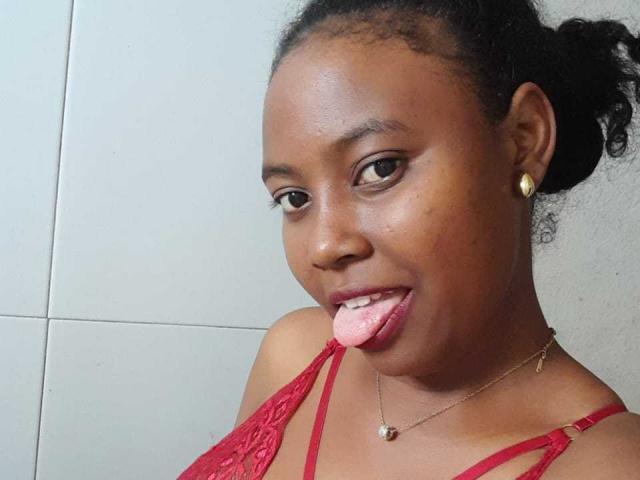 Garbella has dark skin, curly black hair in a bun, red lace bra, gold earrings, necklace, and is sticking out her pink tongue. Her brown eyes are looking at the camera.