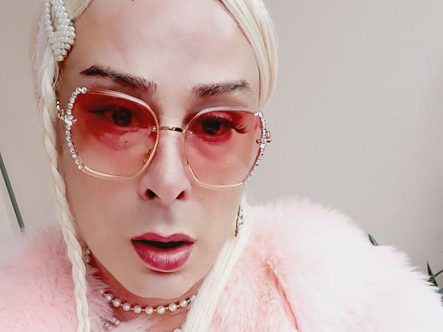 MandyLine with blonde braids, red-tinted glasses, pink fluffy jacket, pearl necklace, and red lipstick, looking surprised. Her nose is small, and her eyes are wide open.