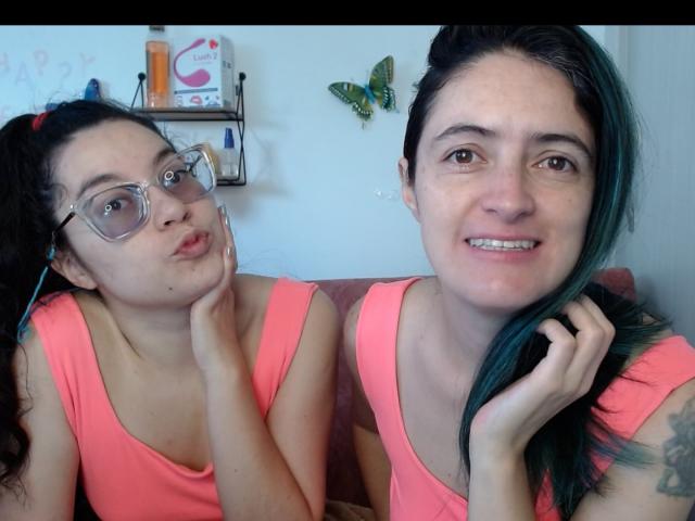 LunaVenus with curly black hair, clear glasses, and pink top, pouting. LunaVenus with green-tinted black hair, pink top, smiling. Both have small breasts, light skin.