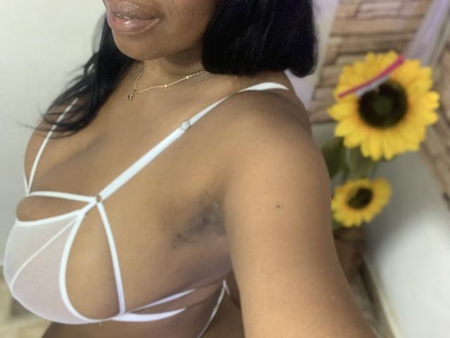 AgataFoxx, à la peau foncée, avec des cheveux noirs, portant un soutien-gorge blanc transparent qui révèle ses seins volumineux et ronds. Des tournesols en arrière-plan, un collier en or et un sourire discret..