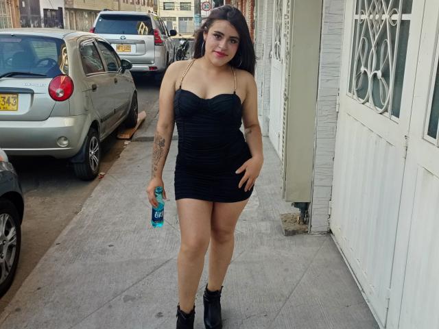 Pamela69s with long dark hair, black strapless mini dress, black boots, holding a blue water bottle, tattoo on right arm, standing on sidewalk, cars in background.