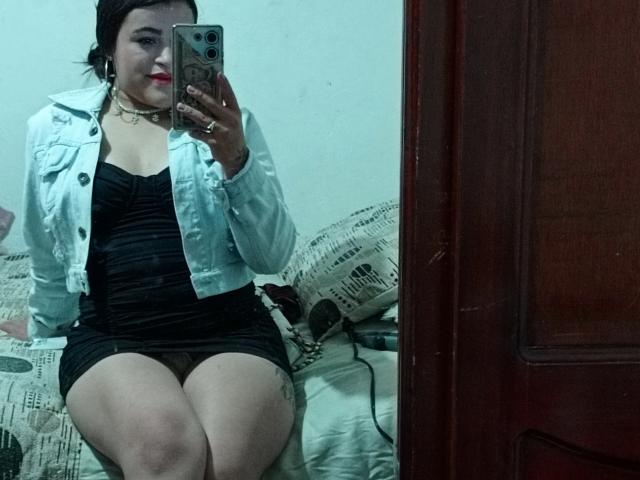 Pamela69s with red lipstick, black dress, and white jacket, sitting on bed, holding phone, showing curvy thighs and tattoo on right leg, in mirror selfie.