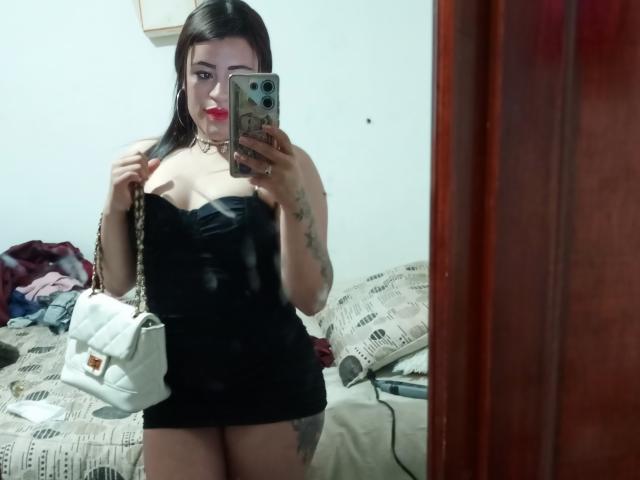 Pamela69s with black strapless dress, red lipstick, holding white purse, tattoos on arm, taking mirror selfie, showing her tits and ass, standing in bedroom, holding phone.