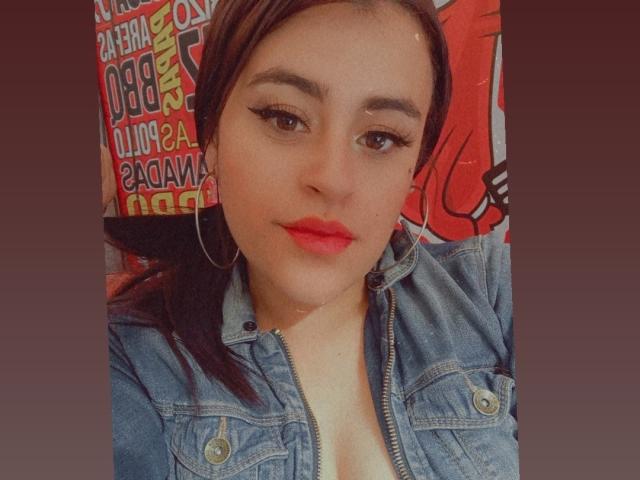 Pamela69s with red lipstick, blue denim jacket, and hoop earrings, showing a little cleavage, has long brown hair and winged eyeliner, looking at the camera.