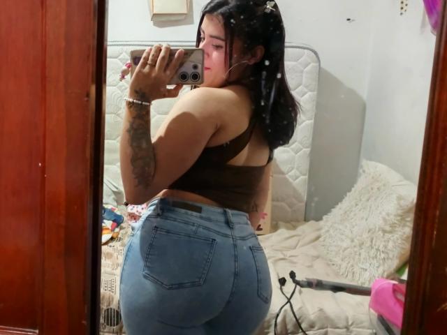 Pamela69s with dark hair, brown tank top, and blue jeans showing her big ass in a mirror selfie, tattoo on arm, holding phone, reflection in wooden-framed mirror.
