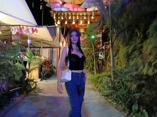 ShantalTurner with dark hair, wearing a black crop top showing her tits and blue jeans, holding a white purse, standing in a nighttime outdoor setting with string lights.