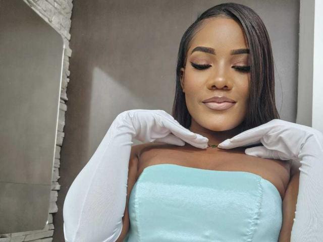 SamiiaSmith with dark brown hair, wearing white gloves and a strapless light blue dress, looking down with a slight smile, showing off her smooth, brown-skinned face and closed eyes.