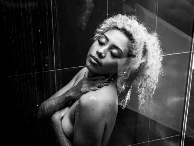 Photo en noir et blanc de LilithWalker, avec des cheveux blonds bouclés, les yeux fermés, une main couvrant ses seins, des gouttes d'eau sur sa peau foncée, et des boucles d'oreilles, appuyée contre un mur carrelé..